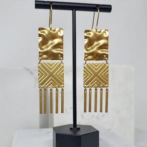 May include: A pair of gold-colored earrings with a hammered texture at the top, a geometric design in the middle, and dangling vertical bars at the bottom. The earrings are displayed on a black stand against a white background.