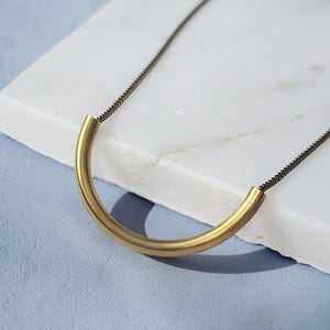 Puede incluir: Un collar que presenta una cadena met&aacute;lica fina y oscura conectada a un colgante de tubo de lat&oacute;n liso, curvado y semicircular. El colgante forma una sencilla figura de arco suspendida de la cadena, creando un elemento de dise&ntilde;o geom&eacute;trico.