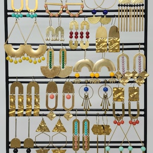 May include: A collection of gold-coloured earrings on a black metal display stand. The earrings showcase a range of designs, including hoops, geometric shapes, and beaded details. The display is set against a neutral backdrop, highlighting the diverse styles.