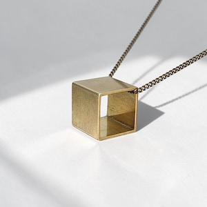 Puede incluir: Un collar de metal en tono dorado con un colgante de cubo hueco suspendido de una cadena fina a juego. El cubo tiene un acabado mate cepillado en sus superficies exteriores y un interior liso y reflectante, creando un diseño geométrico.