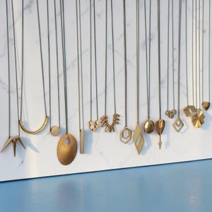 Puede incluir: Una colecci&oacute;n de colgantes de metal dorado colgados de finas cadenas oscuras. Los colgantes presentan diversas formas geom&eacute;tricas, como puntas, una media luna, c&iacute;rculos, un &oacute;valo texturizado, una barra, esferas peque&ntilde;as, hex&aacute;gonos y rombos.