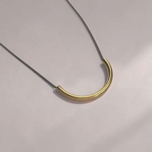 Puede incluir: Un collar que presenta una cadena de metal gris oscuro fina unida a un tubo de metal dorado, liso y curvado en forma de semic&iacute;rculo. La pieza est&aacute; suspendida para crear una silueta limpia en forma de arco con acabados met&aacute;licos contrastantes.