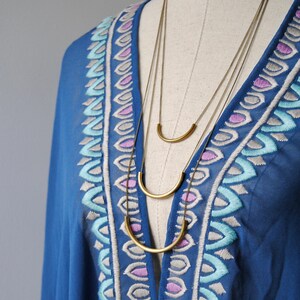 Puede incluir: Un collar dorado de varias capas con tres cadenas finas, cada una adornada con un colgante de barra de metal curvada y lisa. El collar se muestra sobre una prenda azul con escote en V decorado con patrones bordados en azul claro, morado y blanco.