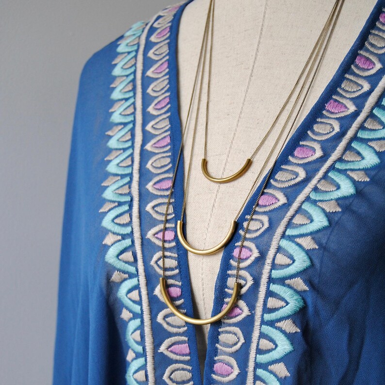 Puede incluir: Un collar dorado de varias capas con tres cadenas finas, cada una adornada con un colgante de barra de metal curvada y lisa. El collar se muestra sobre una prenda azul con escote en V decorado con patrones bordados en azul claro, morado y blanco.