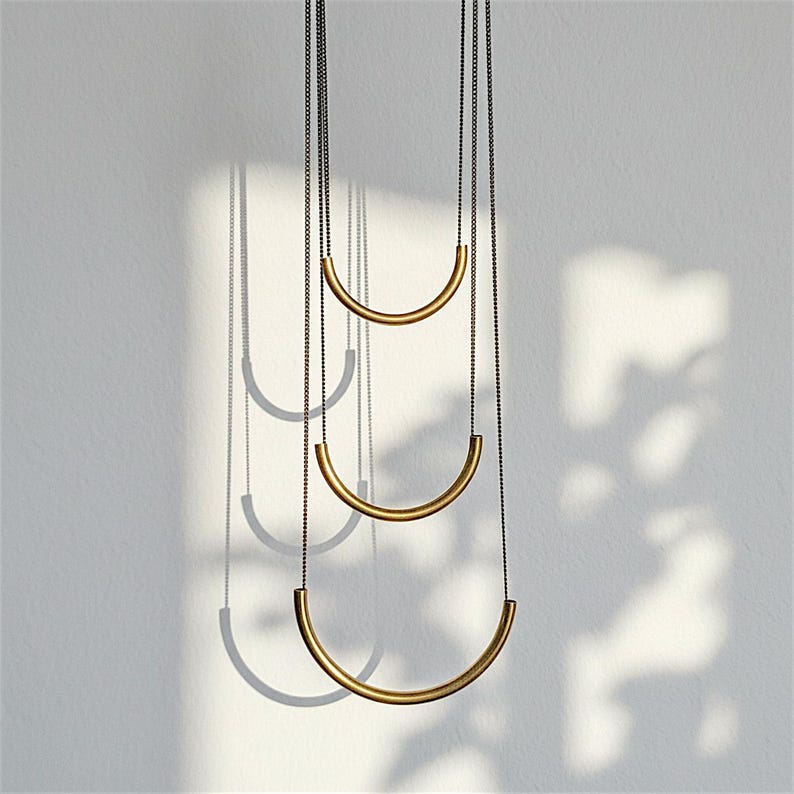 Puede incluir: Un collar de varias cadenas que presenta tres barras met&aacute;licas semicirculares, lisas y de tono dorado, suspendidas de finas cadenas met&aacute;licas oscuras. Las barras est&aacute;n dispuestas en una formaci&oacute;n vertical escalonada, aumentando de tama&ntilde;o de arriba a abajo.