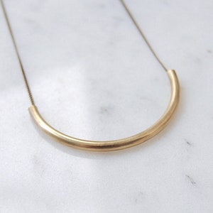 May include: A gold necklace with a simple, curved bar pendant. The pendant is made of a thick, smooth metal and hangs from a delicate chain.