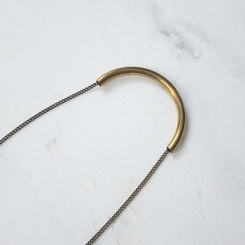 Puede incluir: Un collar que presenta una pieza central de tubo de lat&oacute;n pulido curvado, unida a una cadena de metal fino de tono oscuro. El dise&ntilde;o consiste en una barra de metal lisa en forma de semic&iacute;rculo que se conecta a la cadena delicada en ambos extremos.
