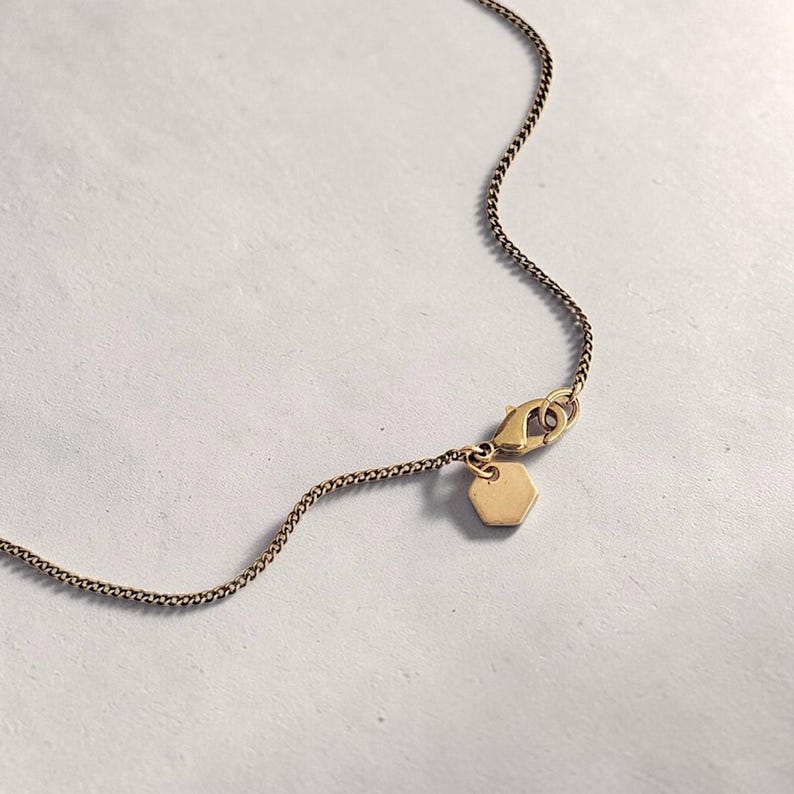 Puede incluir: Un collar de cadena de metal en tono dorado con un cierre de mosquet&oacute;n y un peque&ntilde;o dije hexagonal plano unido al cierre. Los delicados eslabones de la cadena son uniformes en tama&ntilde;o y color, creando una pieza de joyer&iacute;a sencilla y funcional.