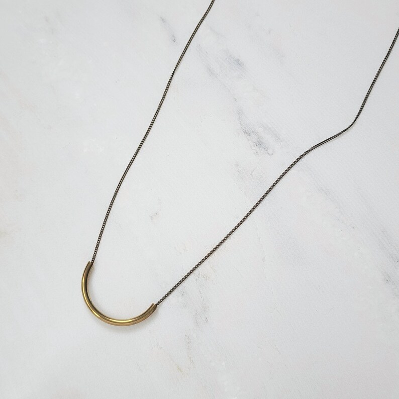 Puede incluir: Un collar que presenta una cadena met&aacute;lica fina y oscura con un colgante de barra de lat&oacute;n liso, curvado en forma de semic&iacute;rculo. El colgante est&aacute; sujeto a la cadena por ambos extremos, creando un dise&ntilde;o geom&eacute;trico simple, limpio y en forma de U.