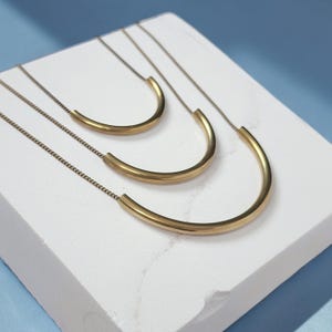Puede incluir: Tres collares dorados, cada uno con un colgante de barra de metal liso, curvo y semicircular suspendido de una cadena delicada. Los collares est&aacute;n dispuestos en una formaci&oacute;n en cascada, con cada colgante aumentando de tama&ntilde;o de arriba a abajo.