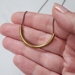 Puede incluir: Un collar que presenta un colgante de tubo de metal dorado, liso y curvado, suspendido de una delicada cadena de metal oscuro. El colgante forma una sencilla figura de arco y la cadena se conecta a ambos extremos del tubo, creando un dise&ntilde;o lineal.