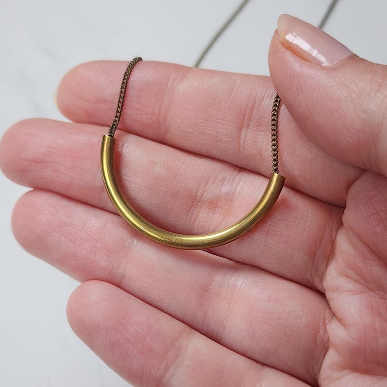 Puede incluir: Un collar que presenta un colgante de tubo de metal dorado, liso y curvado, suspendido de una delicada cadena de metal oscuro. El colgante forma una sencilla figura de arco y la cadena se conecta a ambos extremos del tubo, creando un dise&ntilde;o lineal.