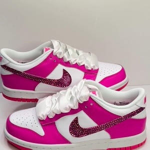 May include: White and hot pink low-top sneakers with a white satin ribbon tied around the laces. The Nike swoosh is covered in pink rhinestones.
