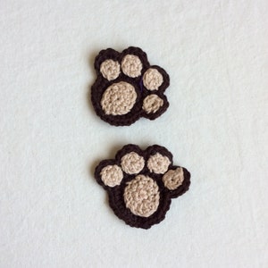 May include: Two crocheted paw print appliques. Each paw print features a dark brown outline with tan-colored pads. The appliques are handmade and suitable for various craft projects, such as embellishing clothing or accessories.