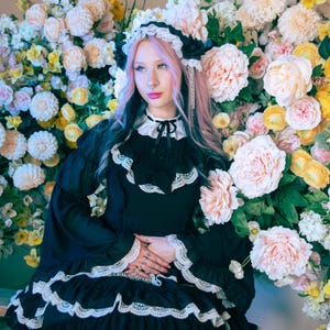 May include: A black dress with white lace trim and a ruffled skirt. The dress has long sleeves and a high collar with a bow. The background is filled with an array of flowers in shades of pink, yellow, and white. Text at the top reads: GLOOMTH & THE CULT OF MELANCHOLY ~ WWW.GLOOMTH.COM