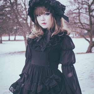 May include: A young person wearing a black dress with lace and ruffles. The dress has a large bow on the front and the person is wearing a black hat with a feather. The person is standing in a snowy landscape.
