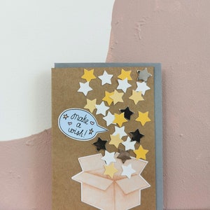 Puede incluir: Tarjeta de felicitación hecha a mano con una parte delantera de papel kraft y un reverso gris. La tarjeta presenta una ilustración de acuarela de una caja abierta con confeti en forma de estrella en amarillo, blanco, negro y plateado. El texto "make a wish!" está en una burbuja.