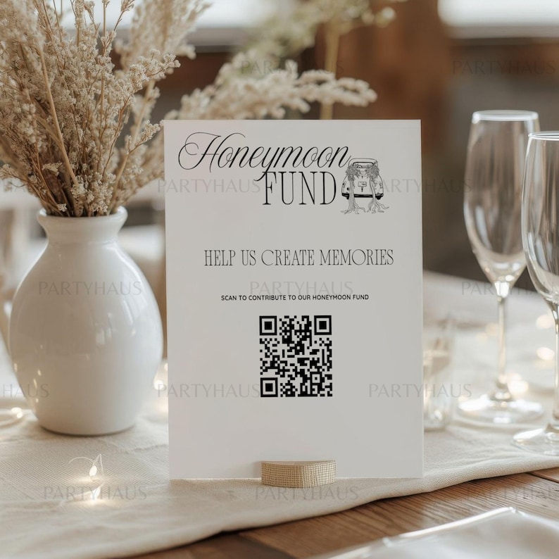 以下が含まれることがあります： 「Honeymoon Fund」と「Help Us Create Memories」という言葉が黒で印刷された白いサイン。テキストの下にはQRコードが表示され、中にはカップルが入った瓶の小さなイラストがあります。サインはテーブルの上に立てられています。