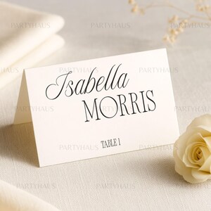 以下が含まれることがあります： 黒い筆記体とセリフフォントで「Isabella Morris」と印刷されたアイボリーの席札。テーブル番号「1」は名前の下に印刷されています。白いバラが右下にあります。
