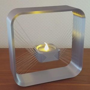 Könnte beinhalten: Ein moderner, silberfarbener Kerzenhalter mit quadratischem Rahmen. Im Inneren hält ein kleiner, quadratischer Kerzenhalter eine brennende, gelbe Kerze. Feine, weiße Linien strahlen vom inneren Kerzenhalter zum Rahmen aus.