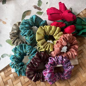May include: A set of seven colorful hair scrunchies made from fabric with a tie-dye pattern. The scrunchies are arranged in a circle on a woven surface.