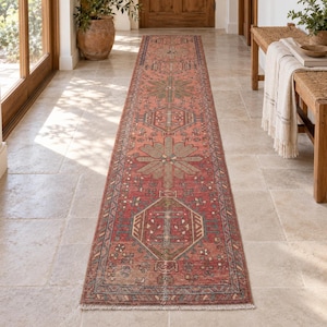 May include: A long, rectangular runner rug featuring a vibrant red base color with intricate geometric and floral patterns in shades of blue, green, and cream. The rug has a symmetrical design with repeating motifs and a fringed edge at the ends.