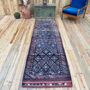May include: A long, rectangular runner rug featuring a dark blue base with a repeating geometric diamond pattern in shades of pink, light blue, and cream. The rug has a fringed edge and a dark border with a small, repeating triangular motif.