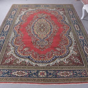 May include: A large, rectangular rug with an elaborate, traditional design. The rug has a central red field with a detailed medallion pattern in beige, blue, and green. A border with floral and geometric motifs frames the central design.