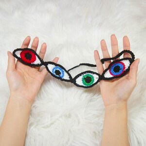 Può includere: Una maschera per gli occhi fatta a mano all'uncinetto con tre paia di occhi rossi, blu e verdi. Ogni occhio è bordato di nero con sfondo bianco. La maschera è esposta su una superficie bianca e soffice.