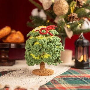 Op de afbeelding: Een handgemaakte gehaakte kerstboom met groene en gele bladeren, een bruine stam en een rode strik bovenop. De boom staat op een gehaakte basis, met een vage kerstboom en decoraties op de achtergrond.