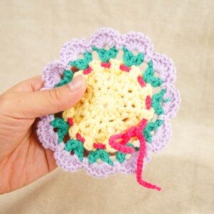 Può includere: Un sottobicchiere fatto a mano, all'uncinetto con un motivo floreale. Il sottobicchiere presenta un centro giallo, circondato da uncinetto verde, rosa e lavanda. Il sottobicchiere ha un diametro di circa 10 cm.