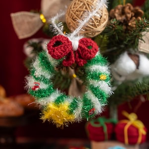 Può includere: Un ornamento a forma di ghirlanda natalizia fatta a mano con un fiocco rosso e una ghirlanda verde, bianca e gialla. La ghirlanda è decorata con piccoli dettagli rossi e verdi ed è appesa a un albero di Natale. L'ornamento è fatto di filo.