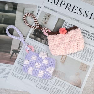 Può includere: Due borsette all'uncinetto fatte a mano in colori pastello. Una è lavanda e bianca, l'altra rosa e marrone. Entrambe hanno motivi intrecciati e fiori decorativi. Sono esposte su uno sfondo di giornale.