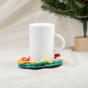 Può includere: Una tazza di ceramica bianca poggia su un sottobicchiere all'uncinetto fatto a mano e colorato. Il sottobicchiere presenta una base verde con accenti rossi, gialli e bianchi. La tazza ha una forma cilindrica semplice e un manico curvo.