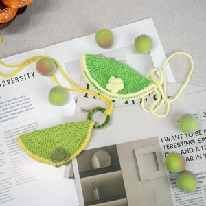 Può includere: Due borsette all'uncinetto a forma di spicchi di lime, in verde brillante con finiture e cinturini gialli. Una borsetta ha un piccolo fiore. Su una superficie bianca con frutta verde sparsa e testo di rivista.