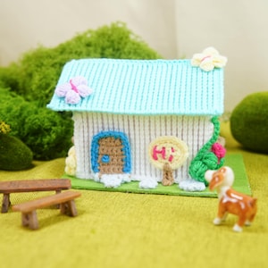 Può includere: Una casa in miniatura fatta a mano con tetto azzurro e pareti bianche. La casa presenta una porta blu, un cartello con la scritta "Hi" e decorazioni floreali. Una piccola panchina marrone e una statuina di cane sono di fronte alla casa.
