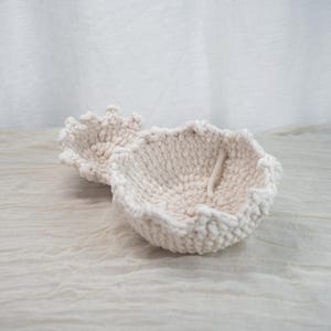 Può includere: Due ciotole all'uncinetto fatte a mano, bianco sporco. Le ciotole hanno un aspetto a maglia grossa e strutturata con un bordo smerlato. Una ciotola è in primo piano e l'altra leggermente dietro. Le ciotole sono su una superficie chiara e strutturata.