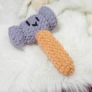 May include: Two handmade dog toys. One is a lavender-colored bone shape with black stitched eyes. The other is a long, orange-colored, rectangular shape. Both toys are made of a soft, textured material and are laying on a white, fluffy surface.