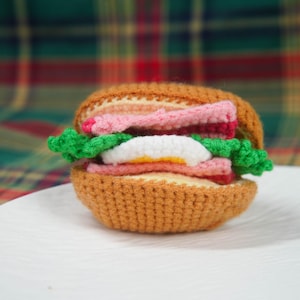 May include: A crocheted sandwich with a brown bun, pink ham, white egg, and green lettuce. The sandwich is sitting on a white plate. The yarn colors are vibrant and the stitches are tight, creating a realistic look.