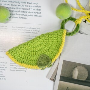 Può includere: Accessorio fatto a mano all'uncinetto a forma di avocado, verde con bordi gialli. L'accessorio ha un piccolo seme rotondo verde ed è collegato a un cordino giallo. Lo sfondo include testo stampato e un frutto verde.