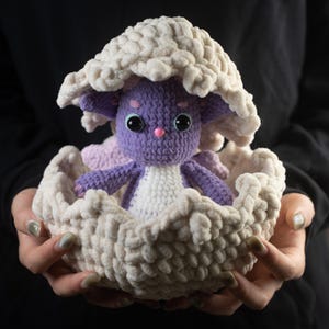 Può includere: Una creatura all'uncinetto fatta a mano in un nido strutturato color crema. La creatura è viola con pancia bianca, dettagli rosa e grandi occhi neri. Il nido ha una parte superiore a forma di fungo, creando un tema fantastico.