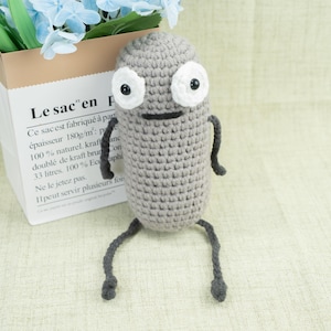Puede incluir: Una muñeca de ganchillo gris hecha a mano con ojos blancos y boca negra. La muñeca tiene brazos y piernas pequeños. La muñeca está frente a una bolsa de papel con el texto "Le sac en" impreso.