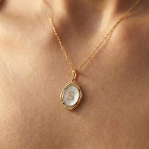 May include: A gold-colored necklace with a pendant featuring a white oval-shaped inlay and the letter "S" in the center. The pendant is framed by a decorative border. The necklace is displayed on a person's skin.