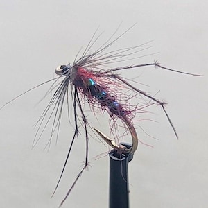 May include: A handmade fly fishing lure. The lure has a gold hook, with a body of red, black, and iridescent fibres. Long, thin, dark brown fibres extend from the body, resembling legs and antennae.