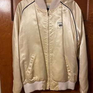 May include: A cream-colored satin bomber jacket with a black zipper and black trim. The jacket has a white ribbed collar and cuffs. The jacket has a Columbia Records logo on the left chest.