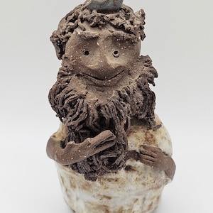May include: A ceramic figurine of a bearded man with a small blue bird perched on his head. The man is smiling and has a textured, brown surface.