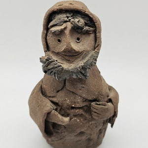 May include: A brown ceramic figurine of a person wearing a hooded cloak. The person has a beard and a smiling face.