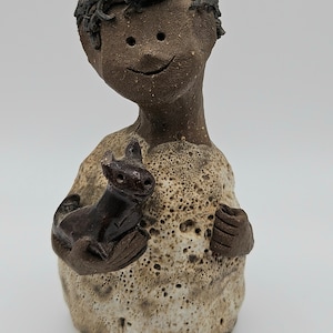 May include: A ceramic figurine of a person with brown skin holding a small brown ceramic animal. The person has a smiling face and is wearing a light brown shirt with a textured surface.