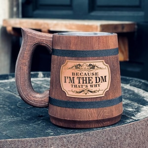 May include: A brown wooden beer stein with a black band around the top and bottom. The stein has a metal plaque with the text "Because I'm the DM That's Why" and a stylized image of a 20-sided die.