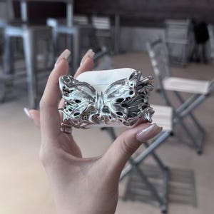 May include: A white wireless earbud charging case featuring a three-dimensional, metallic silver butterfly ornament attached to the front. The butterfly has a liquid, molten-like texture with intricate, flowing wing details that cover the case surface.